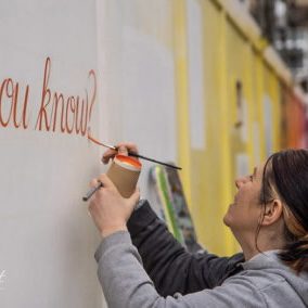 Signwriting the words Did you know? in orange paint onto white hoarding boards as part of a mural collaboration with Wycombe Museum