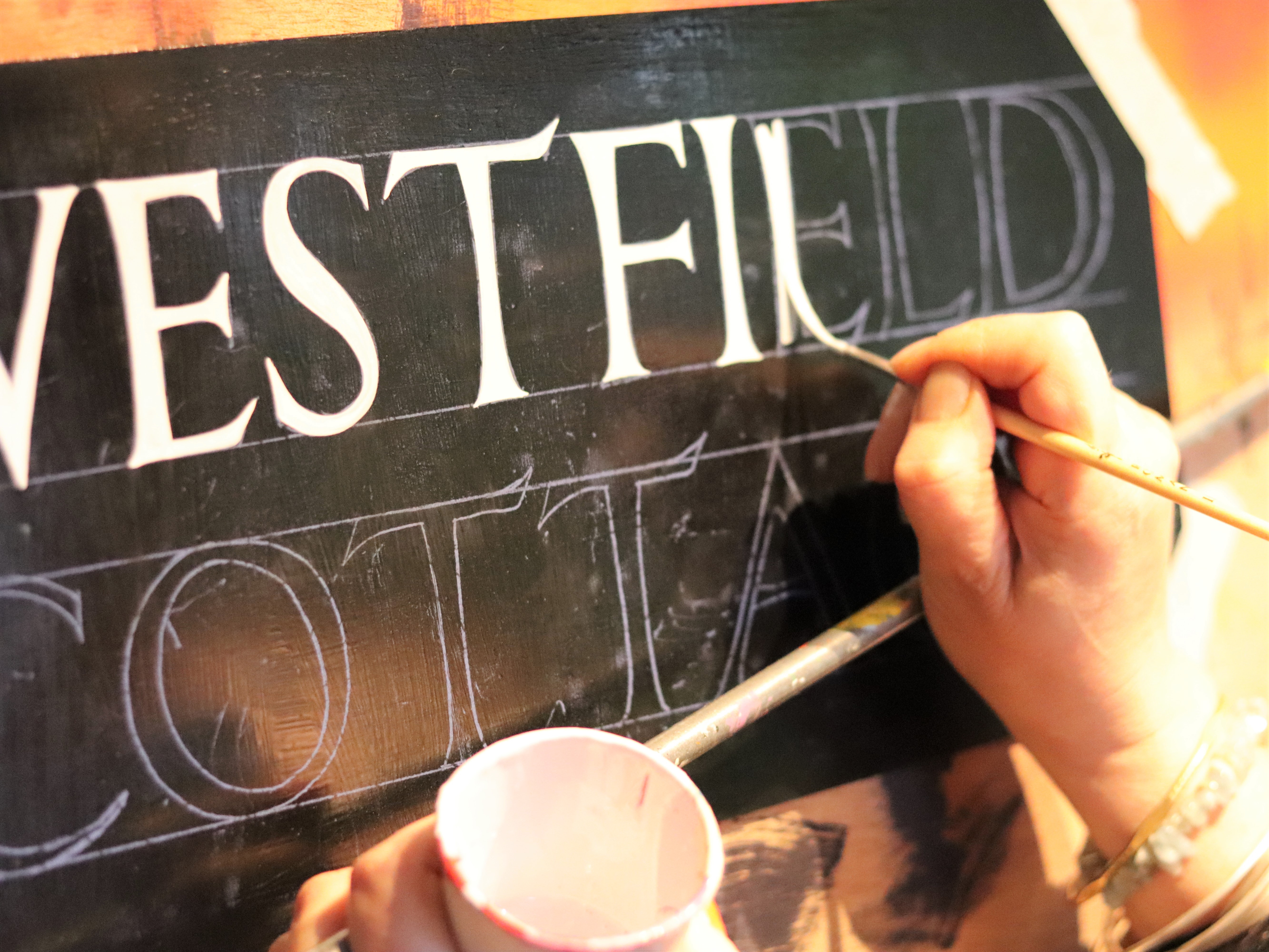 Commission for a bespoke hand-crafted sign showing detail of my hand painting Roman lettering in white 1 shot paint on a black background aluminium sign