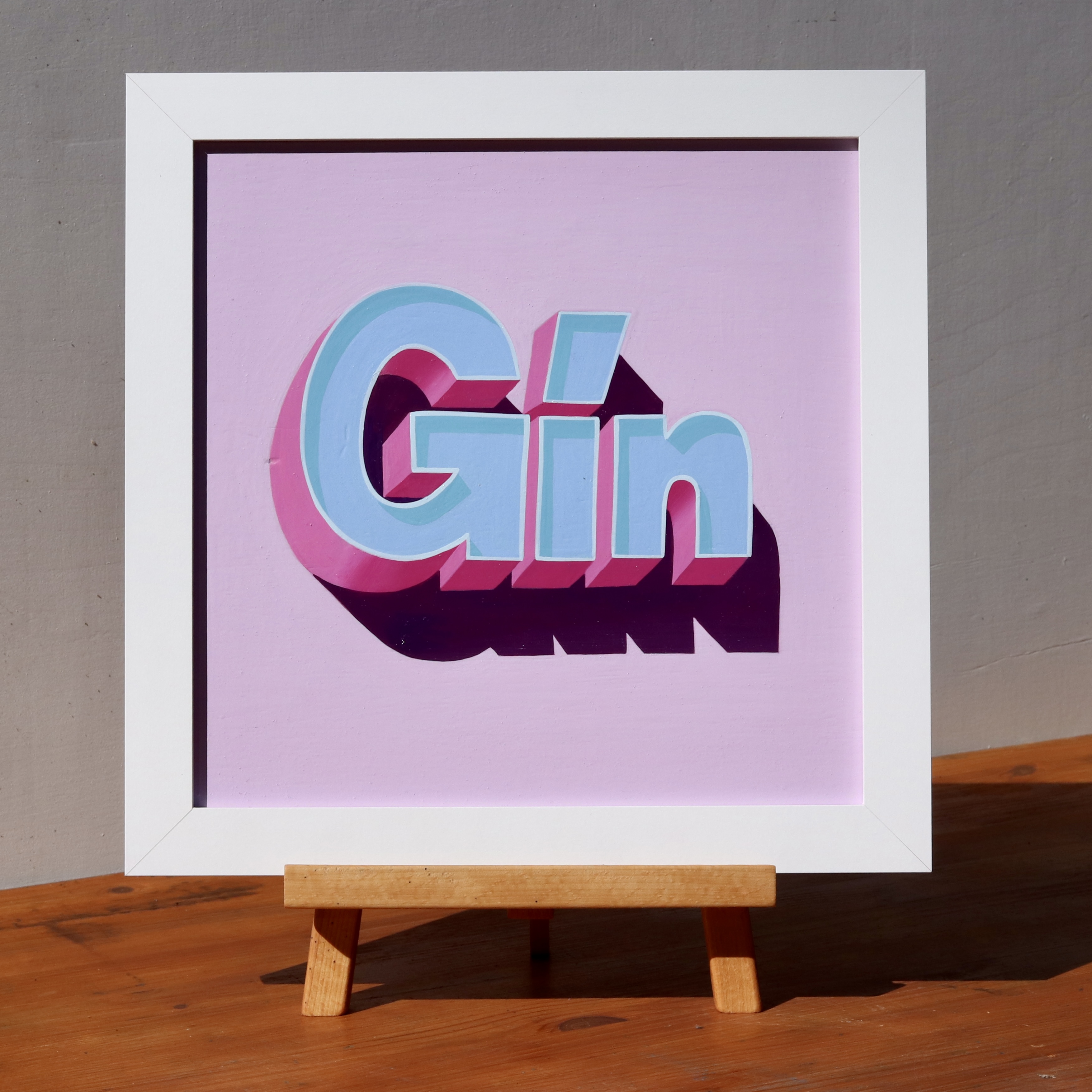 'GIN' hand lettered and painted wall art sign in 3D effect block lettering, light blue and magenta on a lilac background, in a white wooden frame