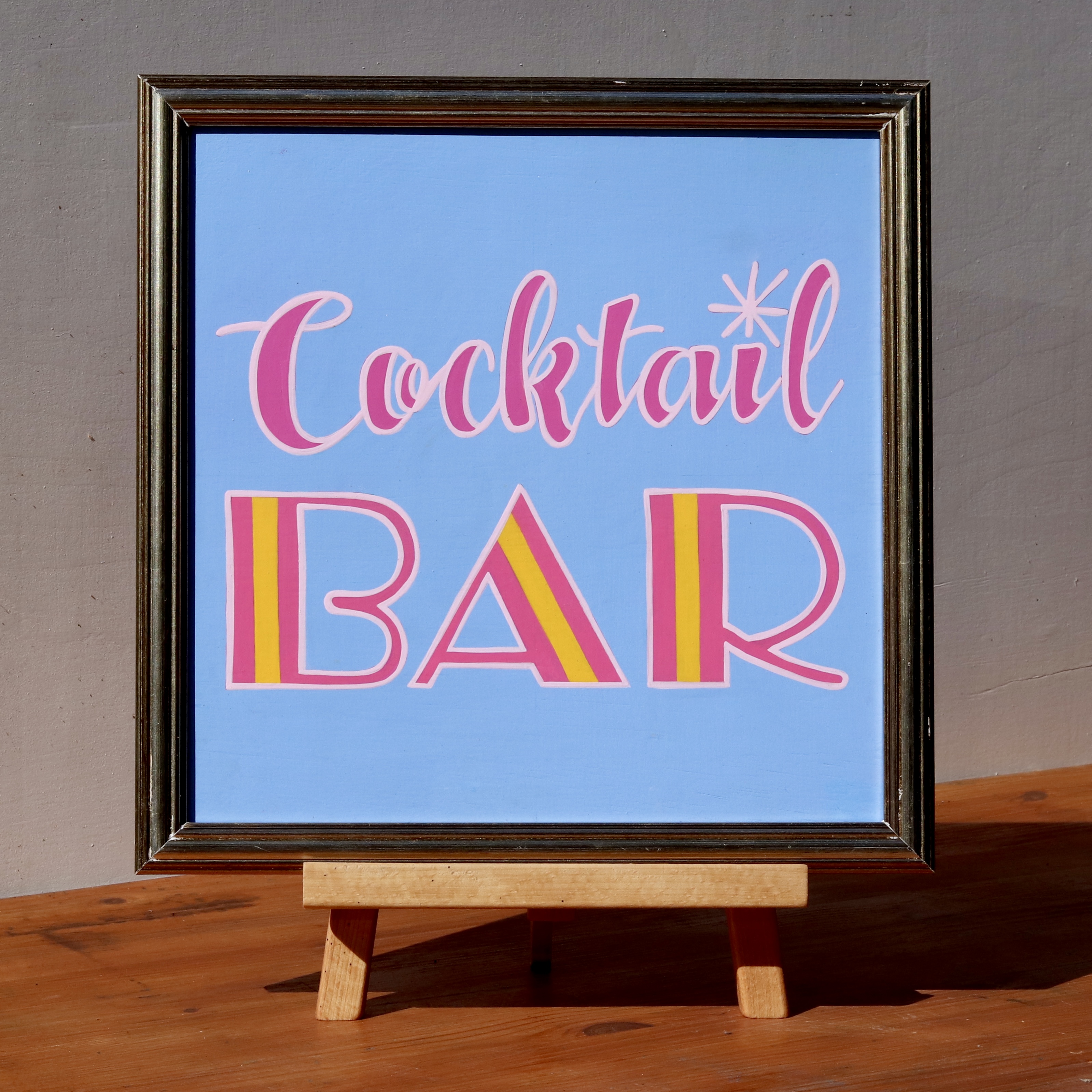 'Cocktail Bar' framed wooden hand-painted word art sign script and art deco style hand lettering in pinks and orange on a light blue background