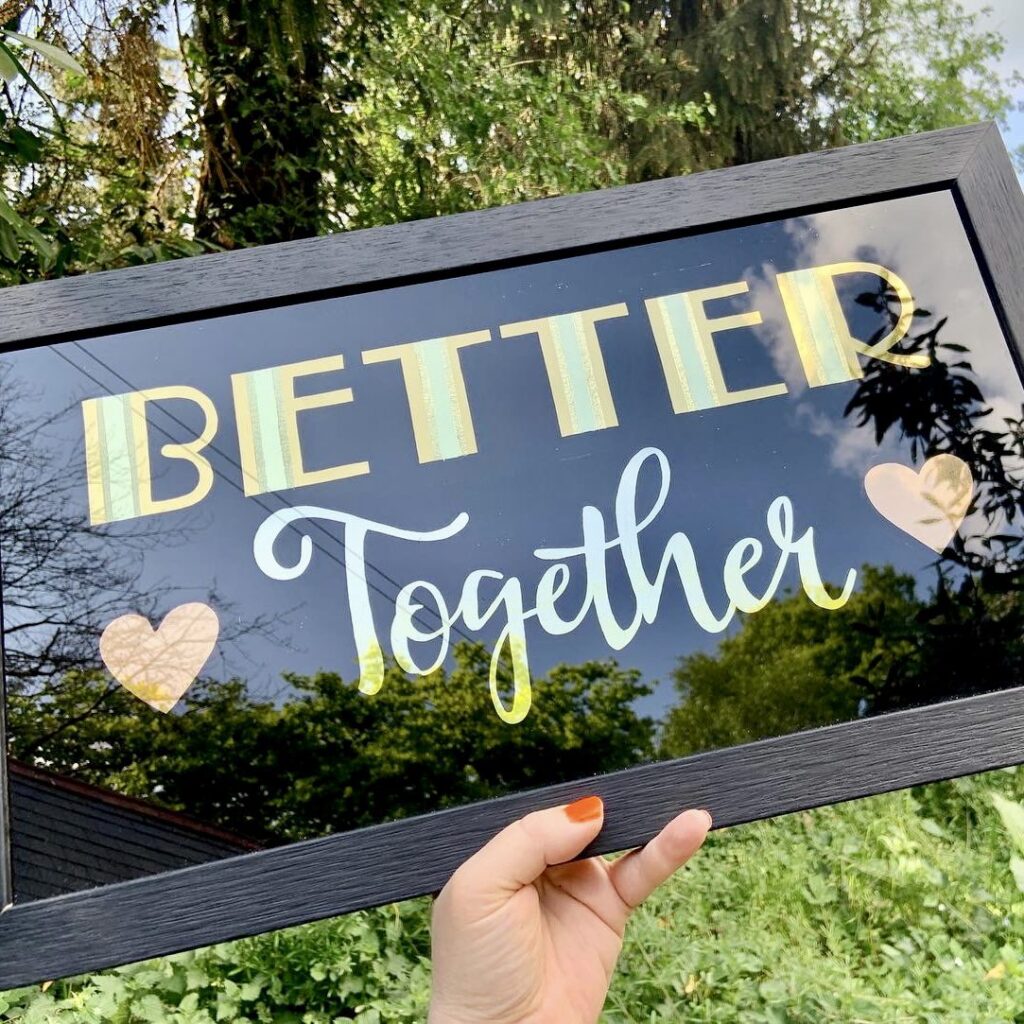 'Better Together' hand lettered typography, gilded onto glass in genuine gold leaf onto glass on a black background with rose gold love hearts on each side