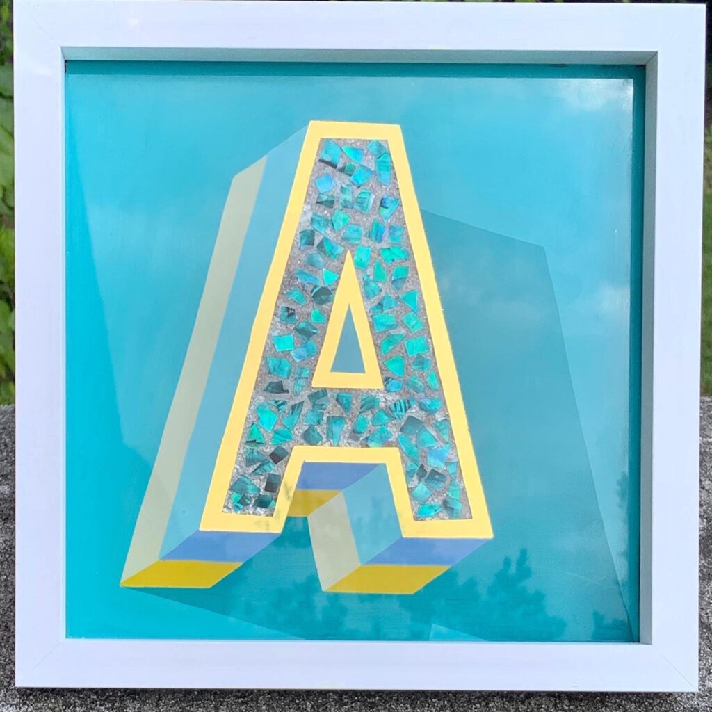 Superstar style letter A in gold leaf, pale blue and yellow block and a cast shadow on a blue background. Hand painted and gilded on glass.