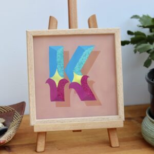 Reverse glass gilded circus style letter K in blues, violets, gold and glitter, finished in an oak wood frame