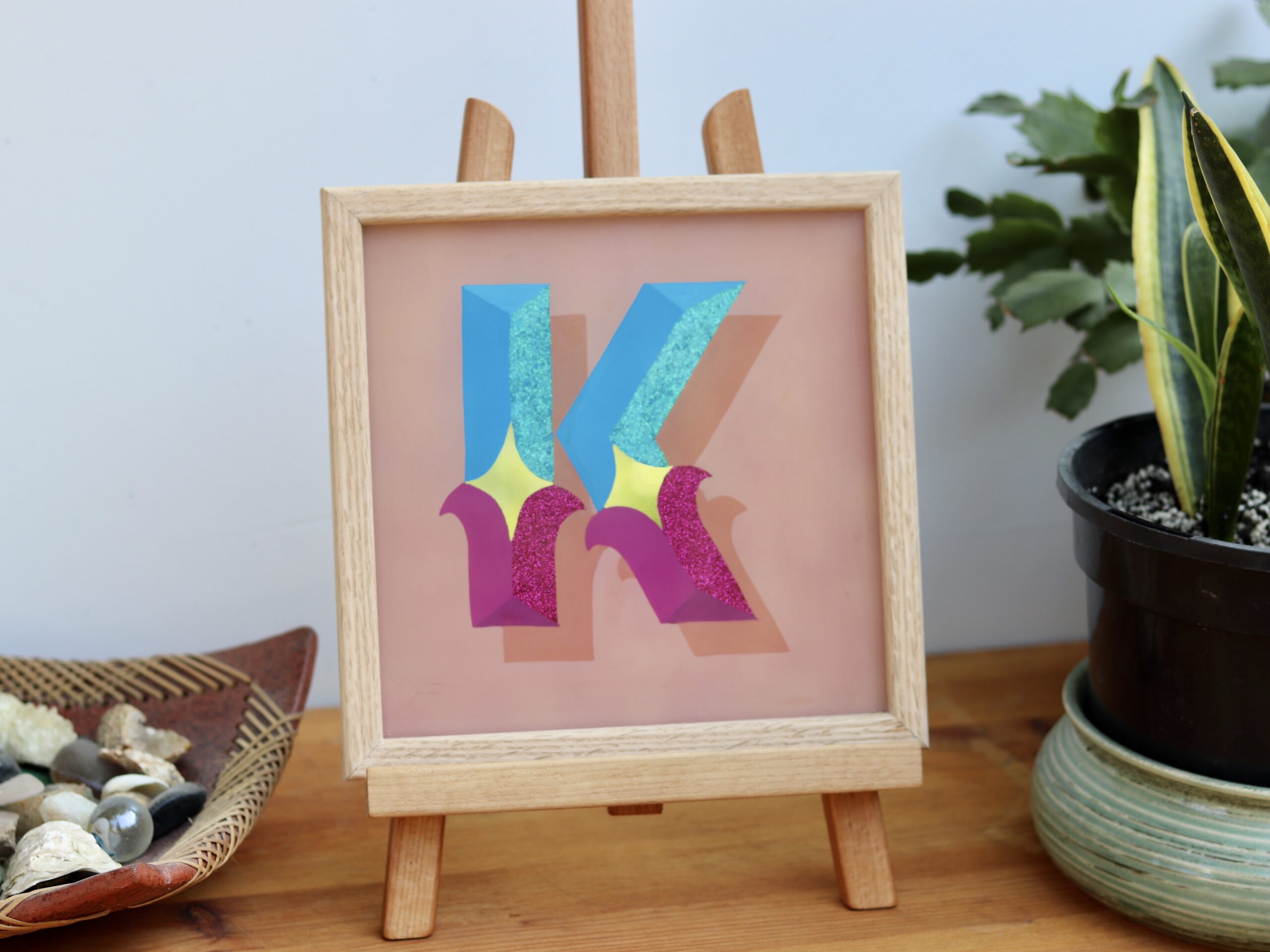 Reverse glass gilded circus style letter K in blues, violets, gold and glitter, finished in an oak wood frame