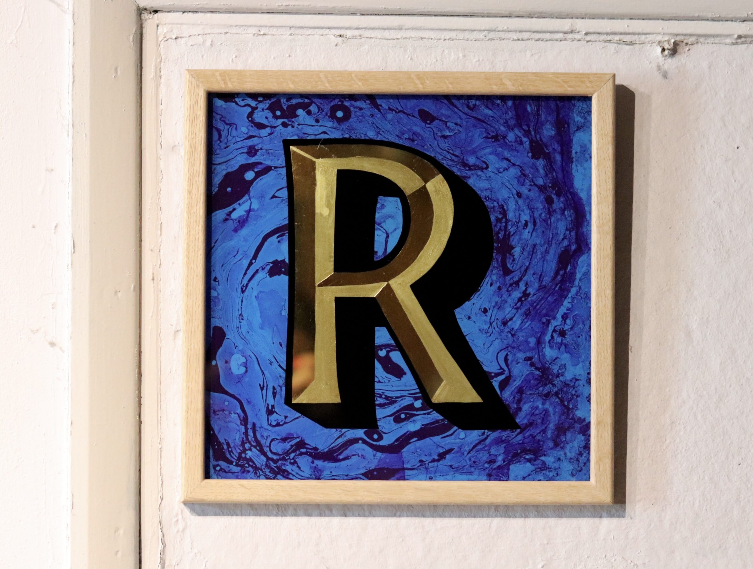 Reverse Glass gilded letter R in 23carat yellow gold and blue marbled backgrounddisplayed on a white wall