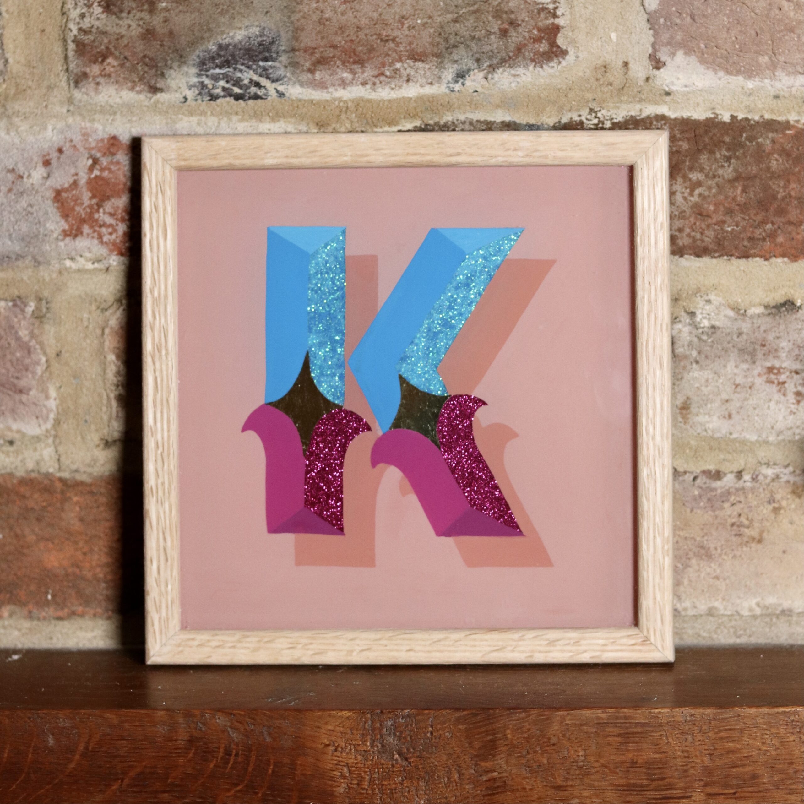 Reverse glass gilded circus style letter K in blues, violets, gold and glitter, finished in an oak wood frame