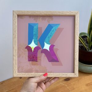 Hand holding a framed reverse glass gilded circus style letter K in blues, violets, gold and glitter