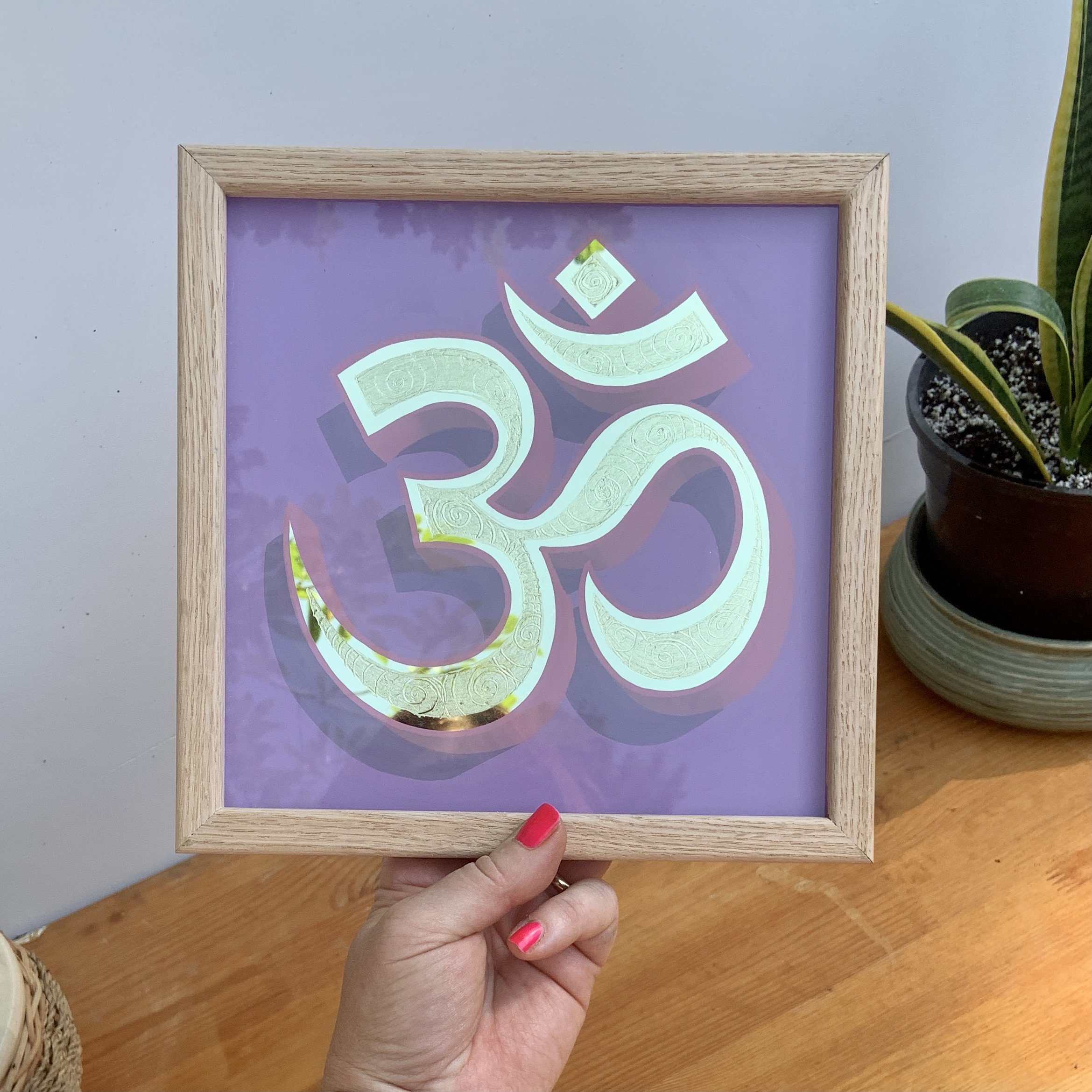 Hand holding an oak framed reverse glass gold leaf Ohm symbol on a light violet background