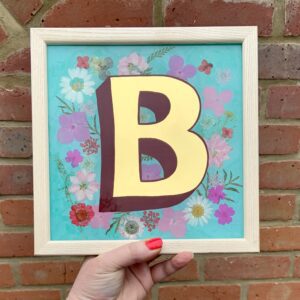 Hand holding a reverse glass gilded letter B surrounded by dried flowers on a light blue background and finished in a light pine wood frame