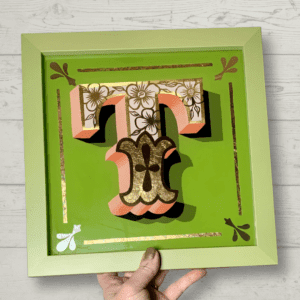 Stunning reverse glass gold leaf circus style letter T handcrafted in 23ct gold leaf with a salmon pink paint blended block on a light green background and framed in olive green wood frame