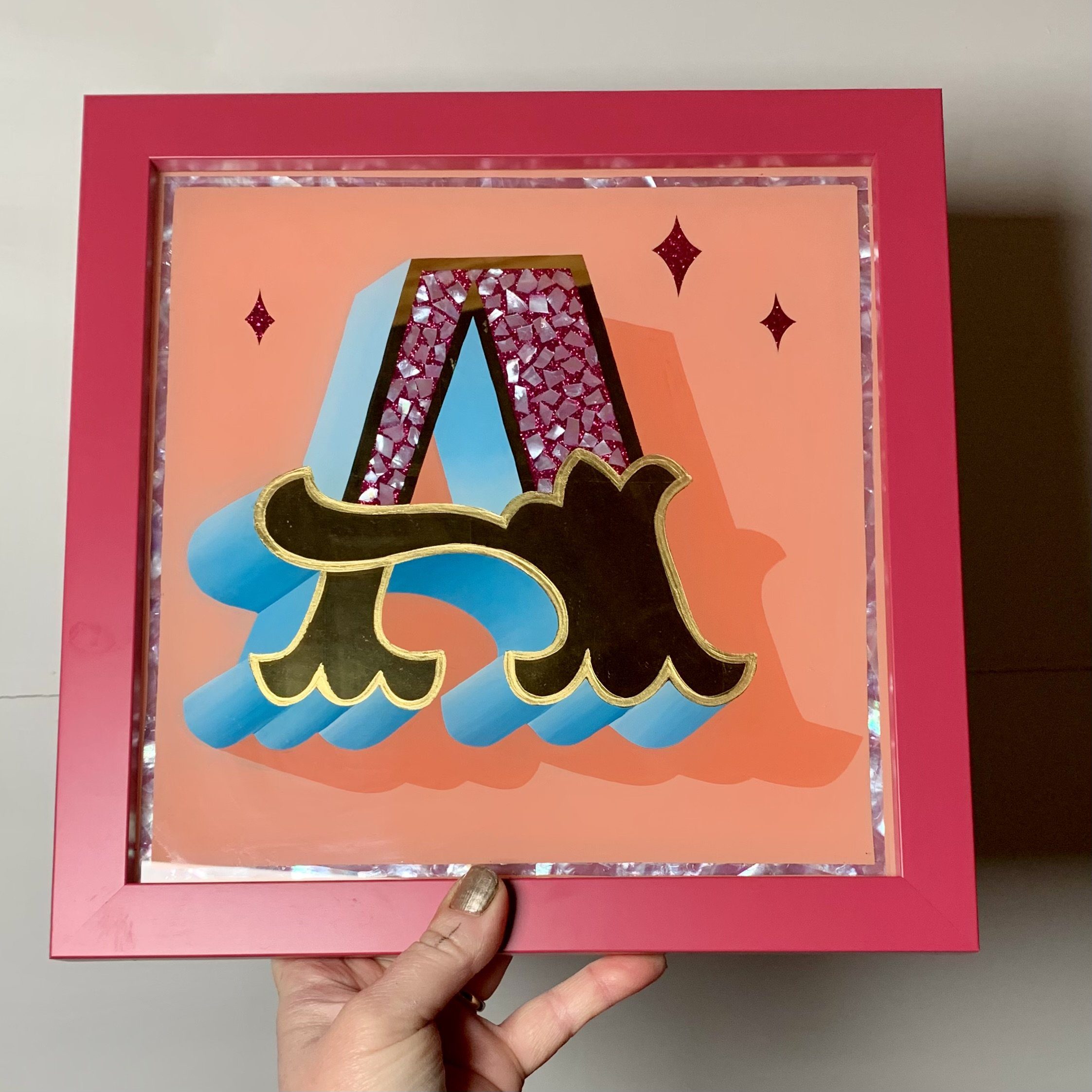 Stunning reverse glass gold leaf circus style letter A handcrafted in 23ct gold leaf with a light blue paint blended block on a salmon pink background and framed in a magenta wood frame