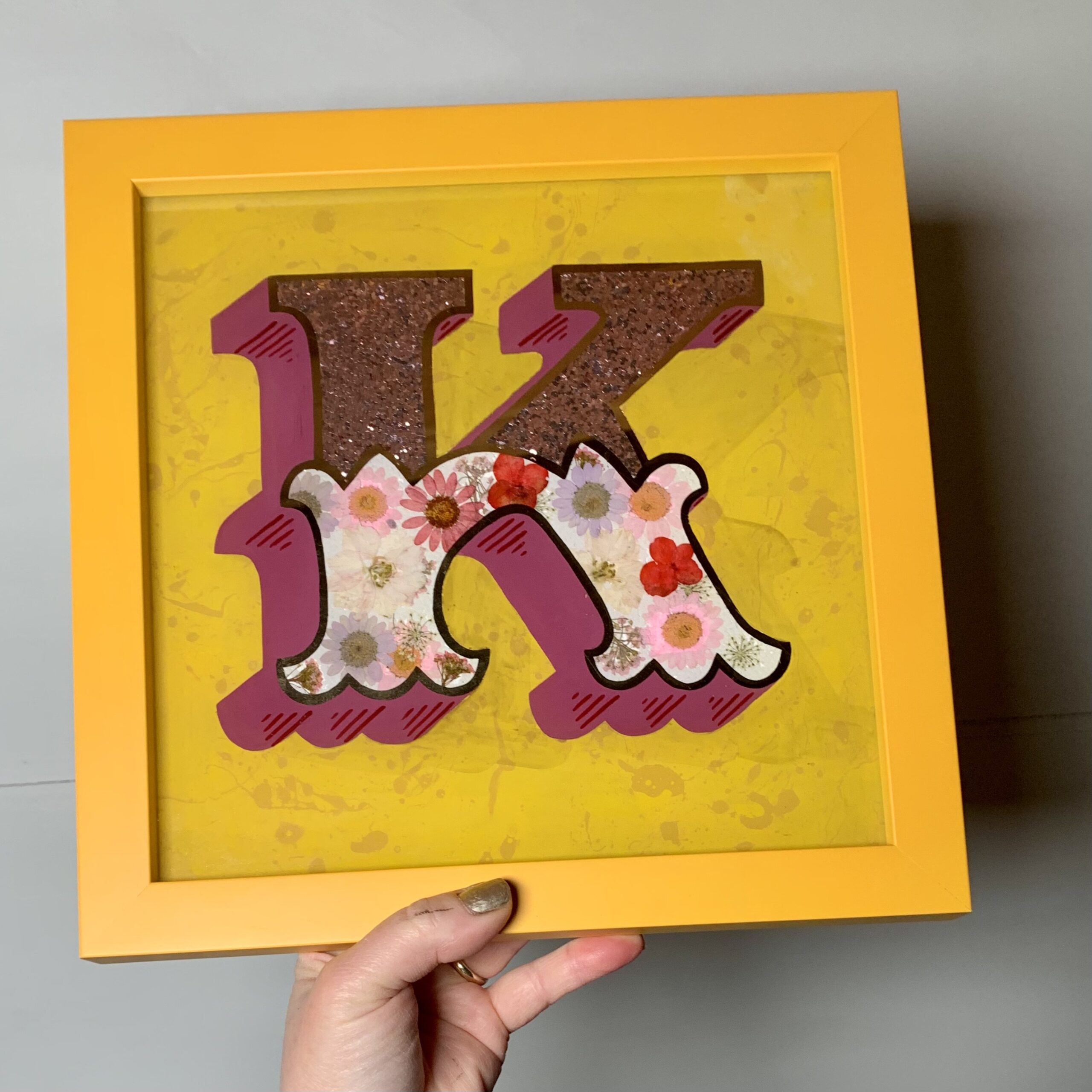 Stunning reverse glass gold leaf circus style letter K handcrafted in 22ct rose gold leaf & infilled with dried flowers and magenta glitter pink set on a marbled paint background and framed in a yellow wood frame