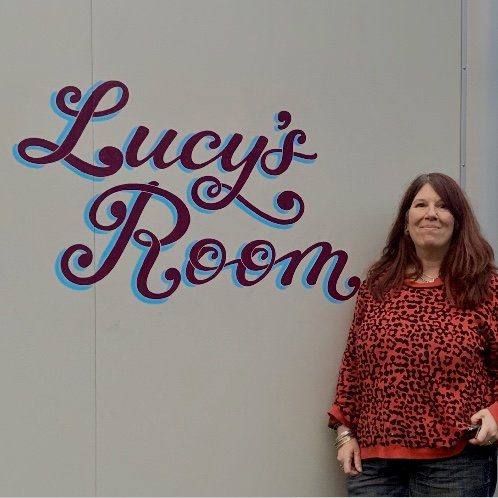 Kat standing next to mural lettering spelling Lucy's Room on the exterior wall of a hospital music therapy room in Oxford