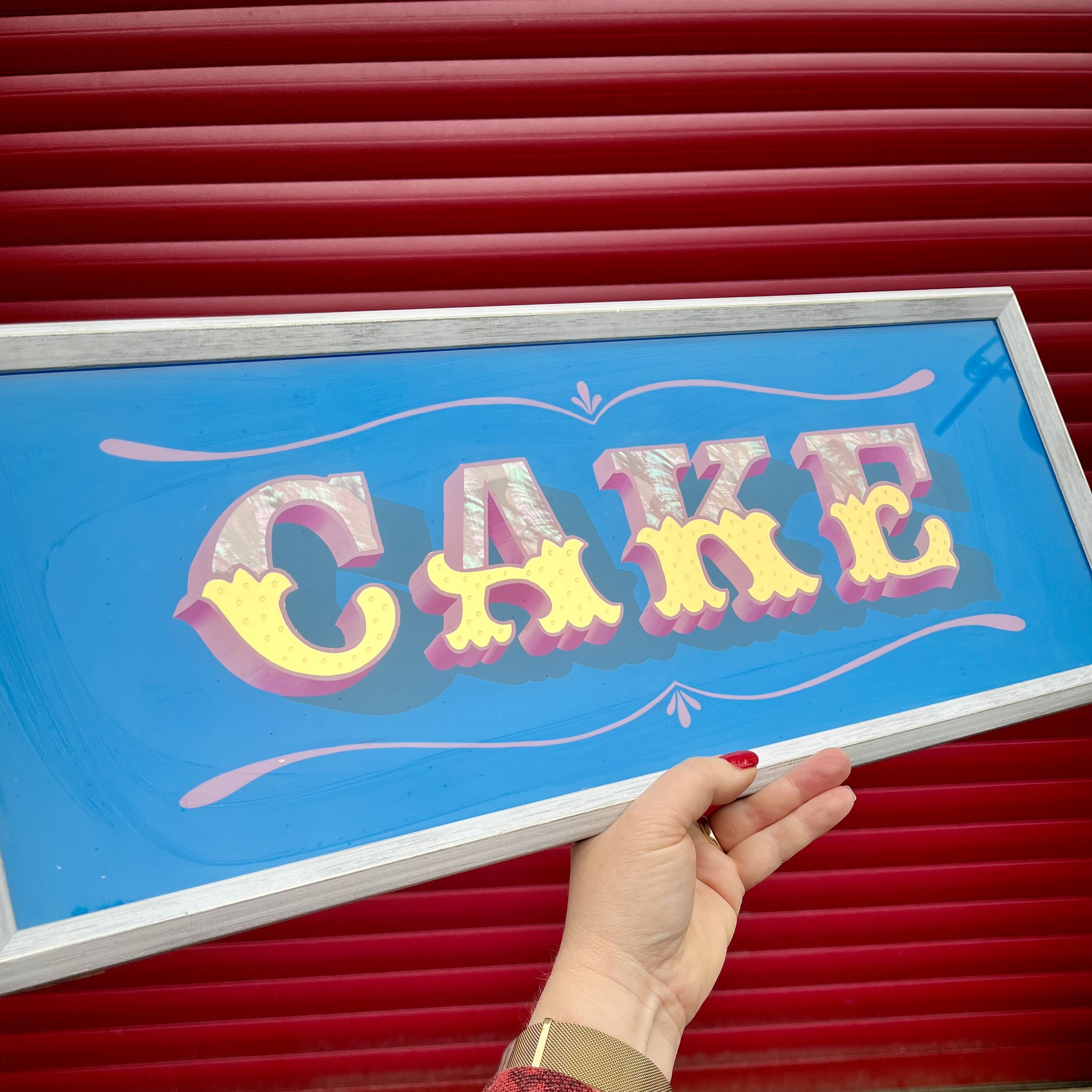 'Cake' Reverse Glass Gold Leaf Wall Art Sign 24CT GOLD LEAF, PINK ABALONE SHELL, MAGENTA AND BLUE enamel paints and yellow gypsy scrolls bordering the word. Finished in a white wood frame