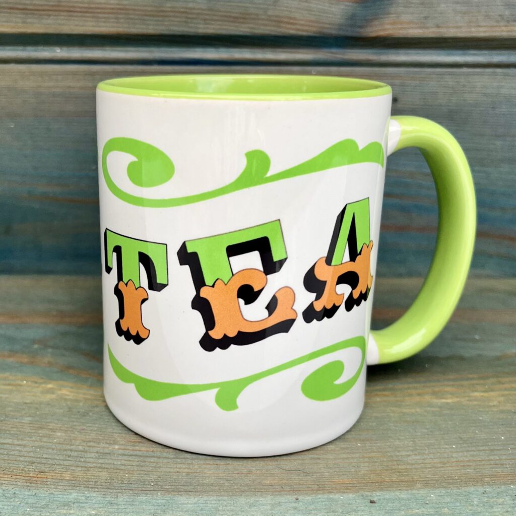 White ceramic 11oz mug with circus style lettering design TEA wrap around and colourful green rim, handle and inside, against a wooden background, side view