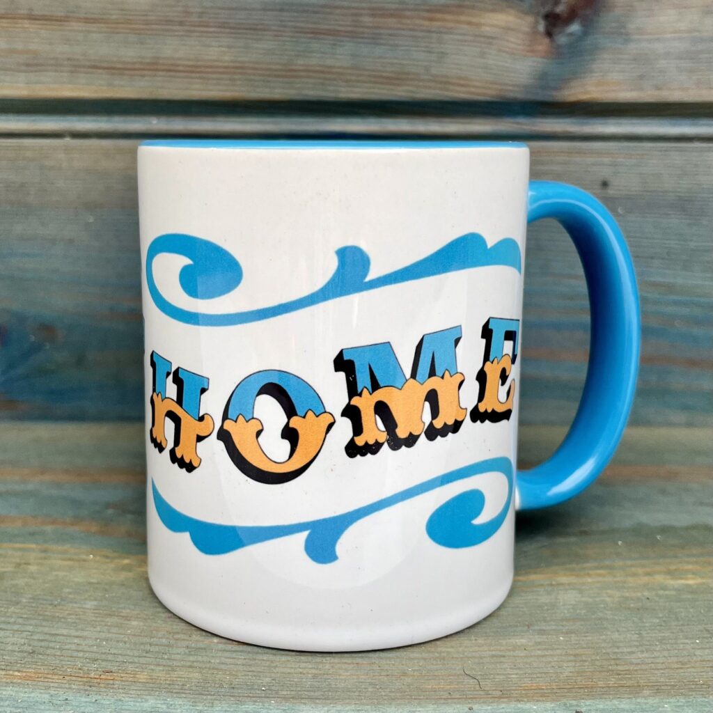 White ceramic 11oz mug with circus style lettering design HOME wrap around and colourful sky blue rim, handle and inside, against a wooden background, side view