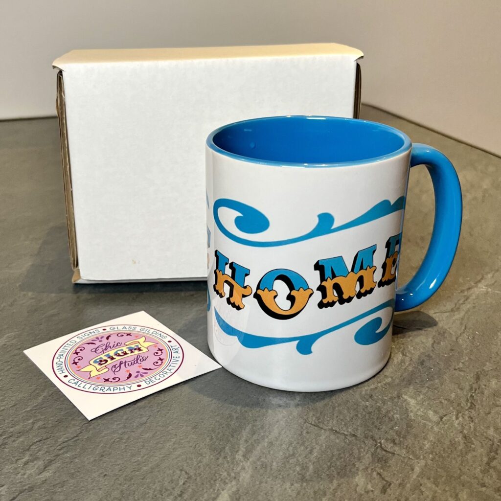 White & blue ceramic mug with Circus Style hand-lettered ‘HOME’ design on a table with postage box and business card