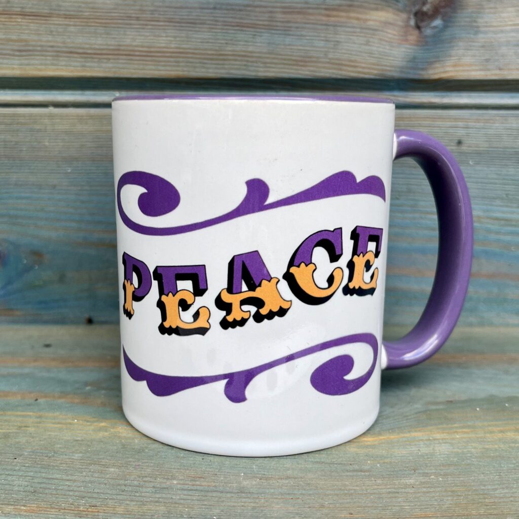 White ceramic 11oz mug with circus style lettering design PEACE wrap around and colourful purple rim, handle and inside, against a wooden background, side view