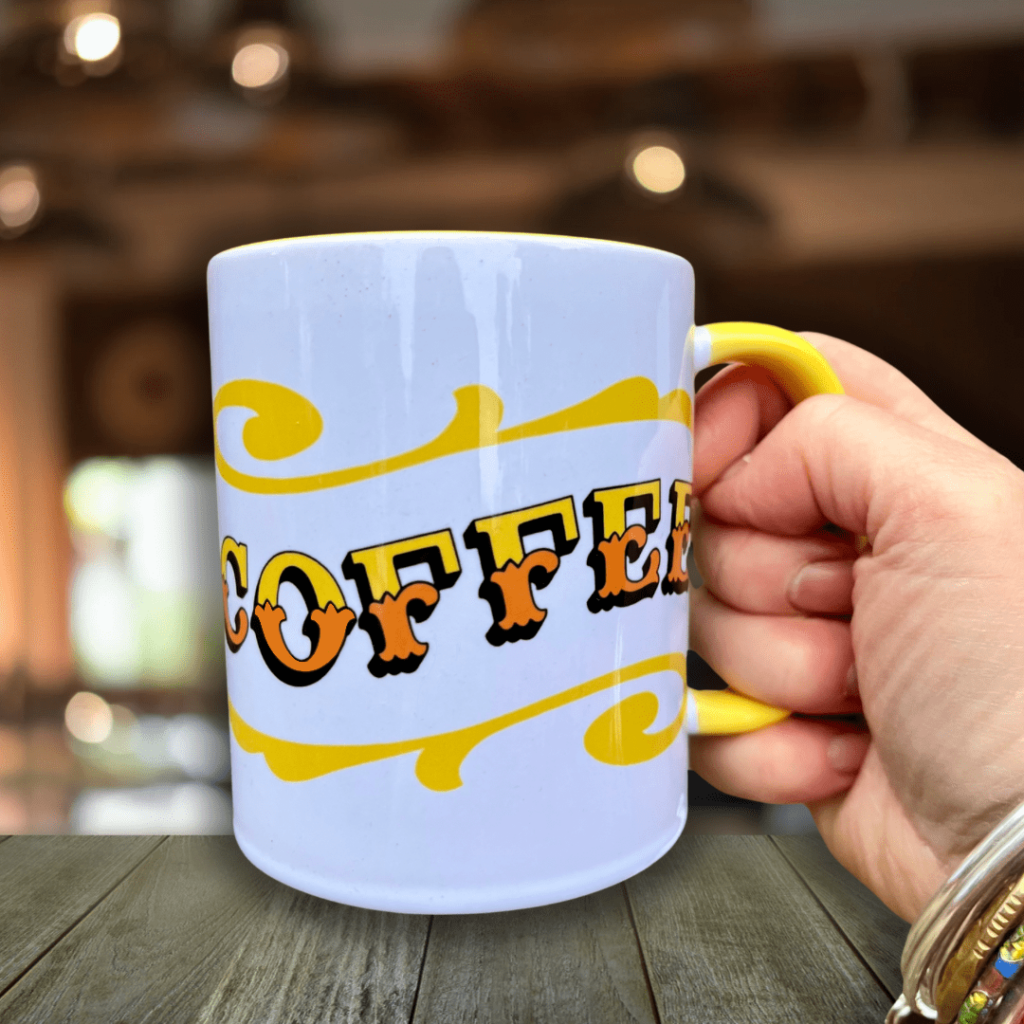 Hand holding Circus Style hand-lettered ‘COFFEE’ Mug with colour inside againt a wooden table in a bar background