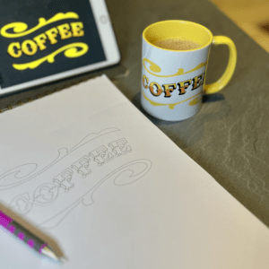 White & yellow ceramic mug with Circus Style hand-lettered ‘COFFEE’ design on a table with the original drawing of the design and a digitised version on procreate ipad