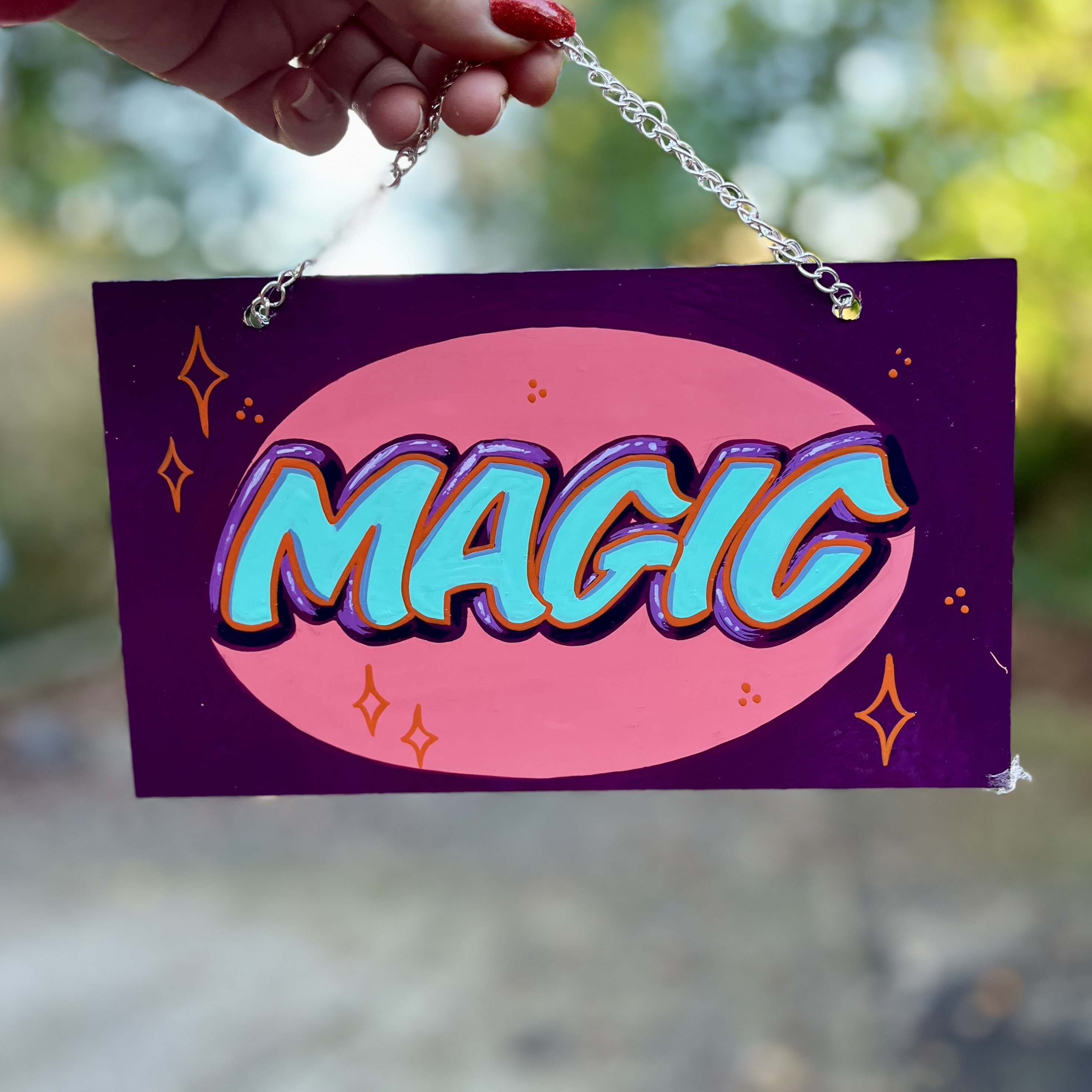 Hand holding a small sign by a silver plated chain, featuring the word MAGIC hand-lettered and painted in light teal and purple signwriting enamels onto a small aluminium panel with a pink oval background purple surround and orange starbursts