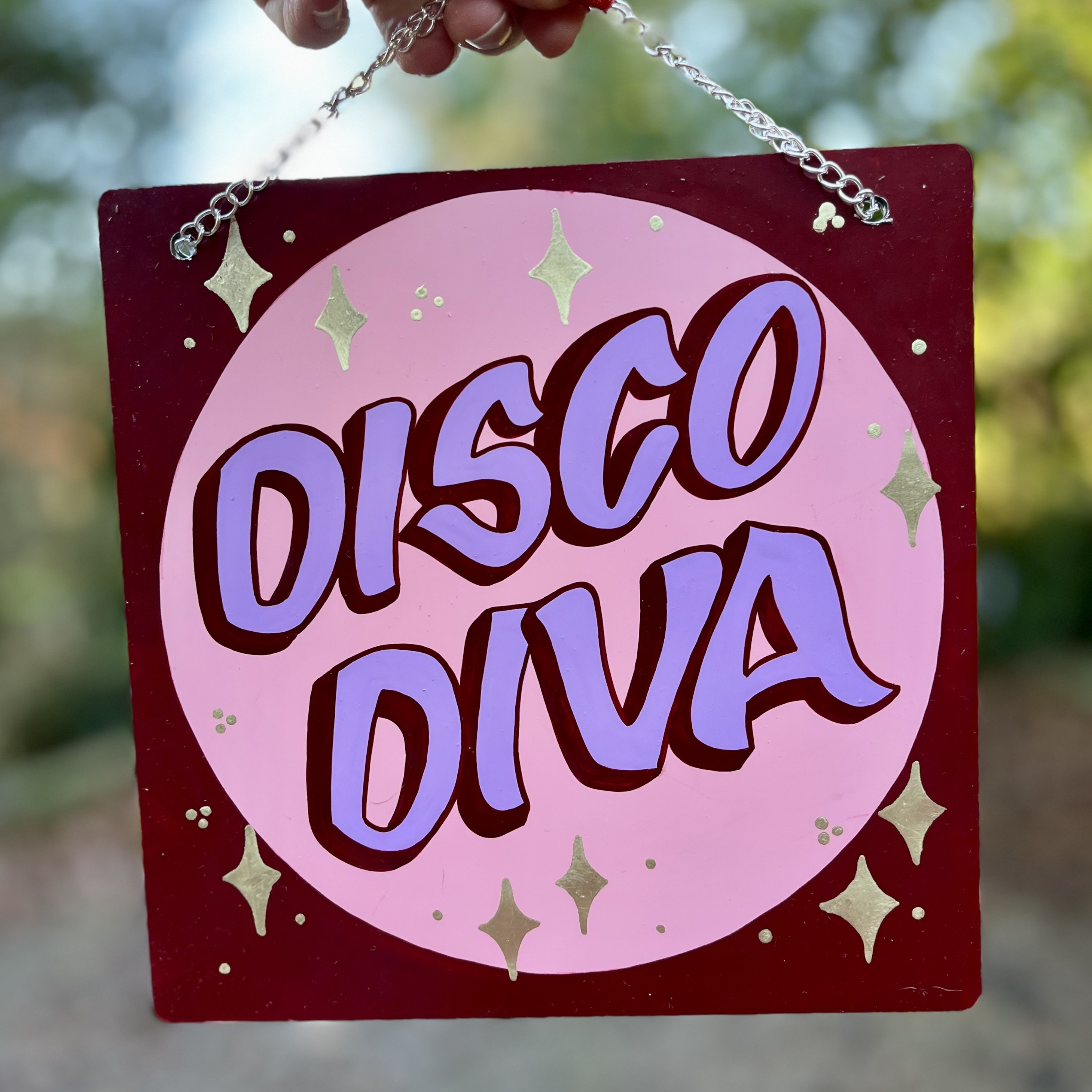 Small hand-painted sign with silver plated hanging chain featuring the words DISCO DIVA in light purple casual style letters outlined in purple against a pink circle burgundy border and gilded starbursts