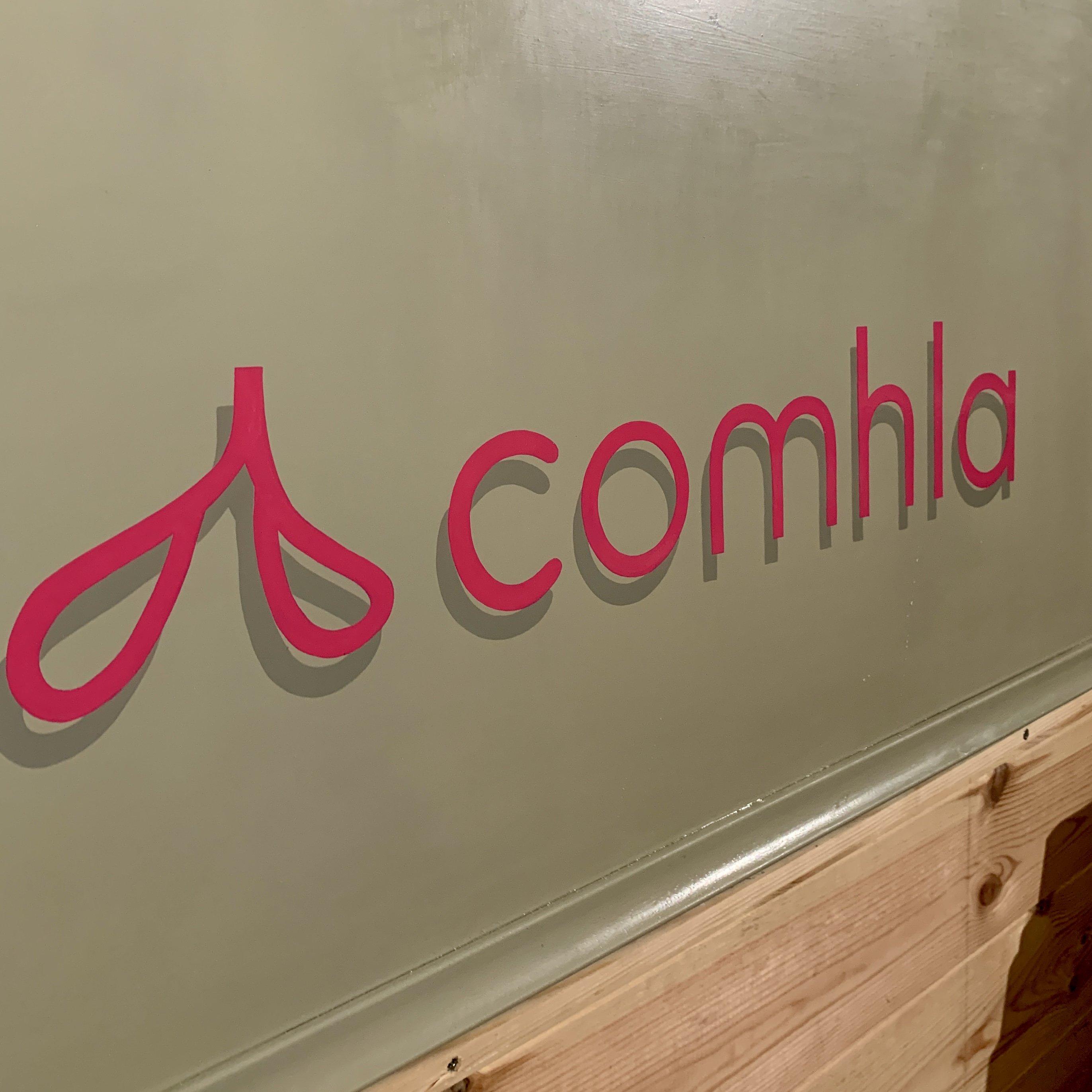 Handpainted replication of logo in shocking pink on a khaki background for Comhla sustainable fashion shop in Long Crendon