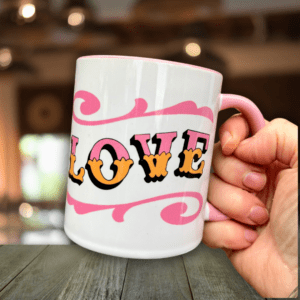 Hand holding white & pink ceramic mug with Circus Style hand-lettered ‘LOVE’ design, over a wooden table in a bar setting