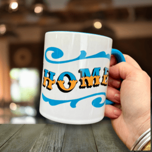 Hand holding white & blue ceramic mug with Circus Style hand-lettered ‘HOME’ design, over a wooden table in a bar setting