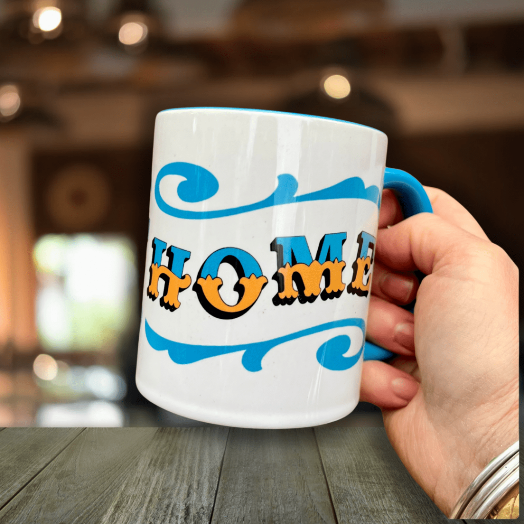 Hand holding white & blue ceramic mug with Circus Style hand-lettered ‘HOME’ design, over a wooden table in a bar setting