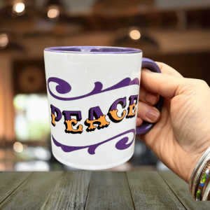 Hand holding white & purple ceramic mug with Circus Style hand-lettered ‘PEACE’ design