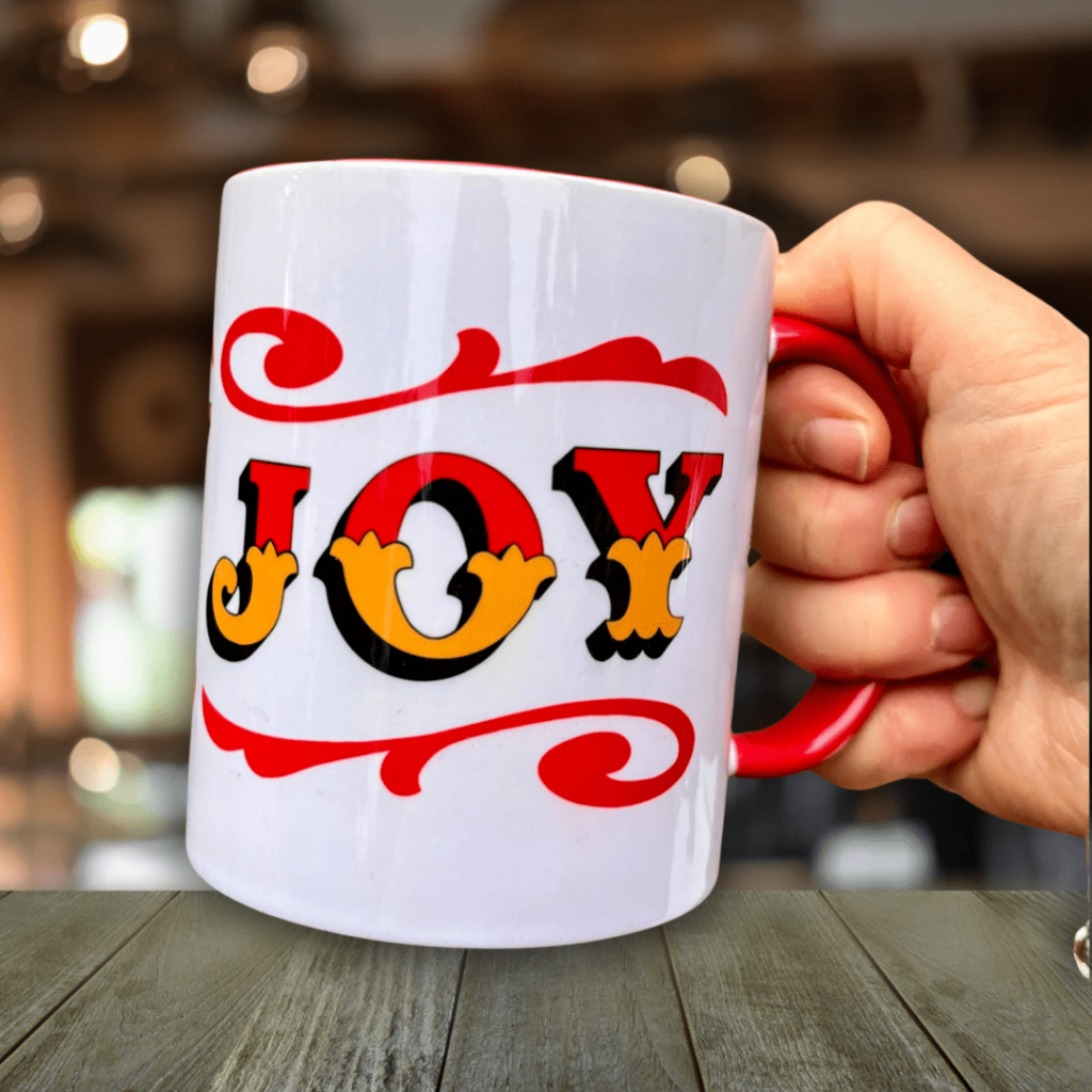 Hand holding white & red ceramic mug with Circus Style hand-lettered ‘JOY’ design, over a wooden table in a bar setting