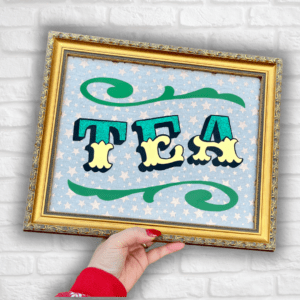 Hand holding 'TEA' in Circus Style Lettering bordered with decorative scrolls, created using real gold leaf, green glitter & green paint and finished in a gold colour carved wood frame in front of a white brick wall