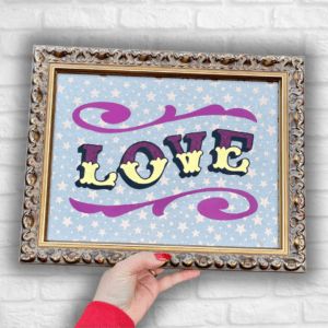 Hand holding 'LOVE' in Circus Style Lettering bordered with decorative scrolls, created using real gold leaf, magenta glitter & magenta paint and finished in a gold colour carved wood frame in front of a white brick wall