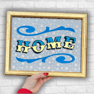 Hand holding 'HOME' in Circus Style Lettering bordered with decorative scrolls, created using real gold leaf, blue glitter & blue paint and finished in a gold colour carved wood frame in front of a white brick wall