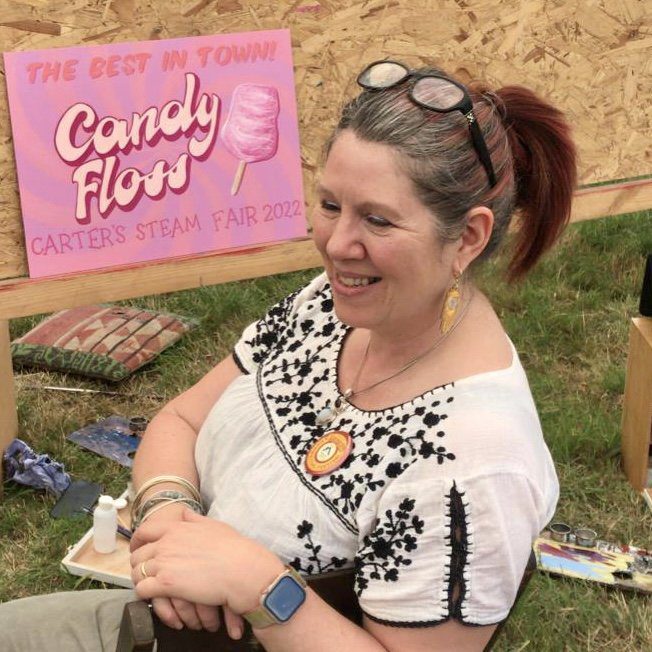 Kat sitting next to an easel with a handpainted sign in pinks and cream for Candy Floss at Carters Stem Fair Signwriting Festival