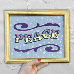 Hand holding 'PEACE' in Circus Style Lettering bordered with decorative scrolls, created using real gold leaf, purple glitter & paint and finished in a gold colour wood frame in front of a white wall