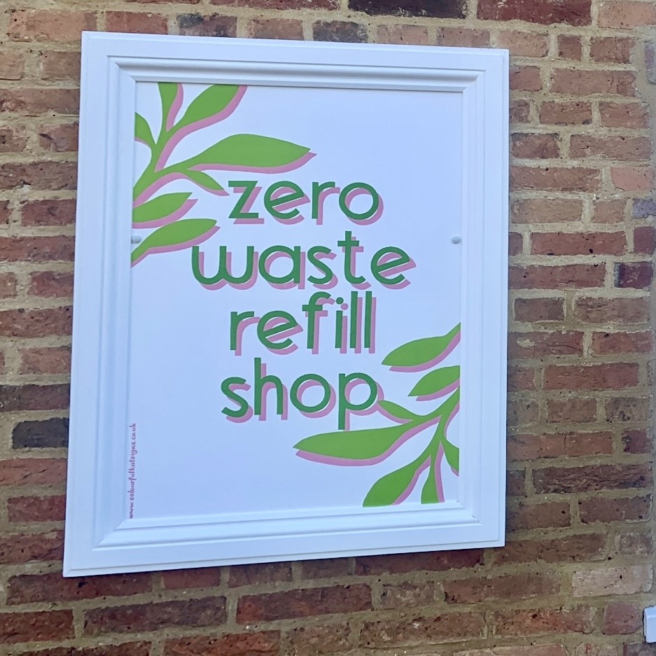 Zero waste refill shop hand-painted shop sign in greens and pink on white background attached to exterior brick walll